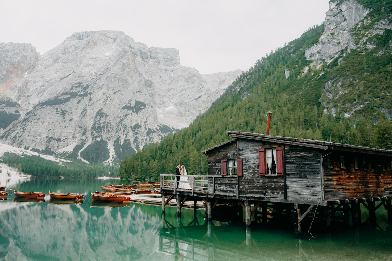 A wooden cabin on stilts sits over a calm lake with several rowboats nearby, surrounded by pine trees and mountains in the background.