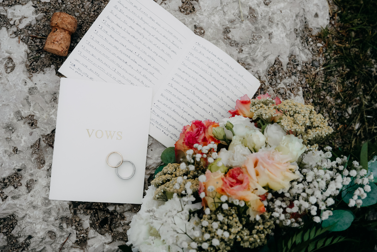 An open book, a white envelope with two rings on top, and a bouquet of flowers are arranged on a stone surface.