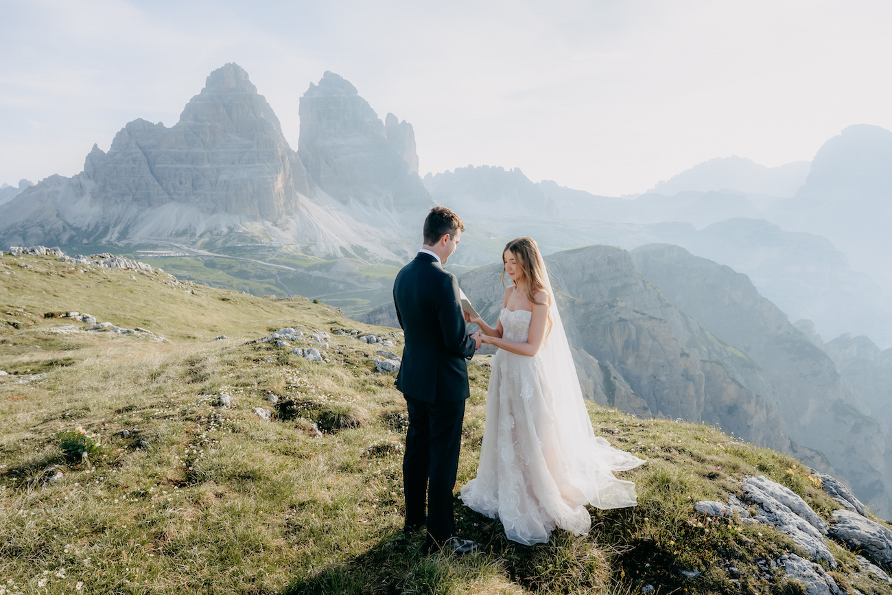 A bride and groom stand facing each other outdoors on a grassy hilltop with rocky mountains in the background.
