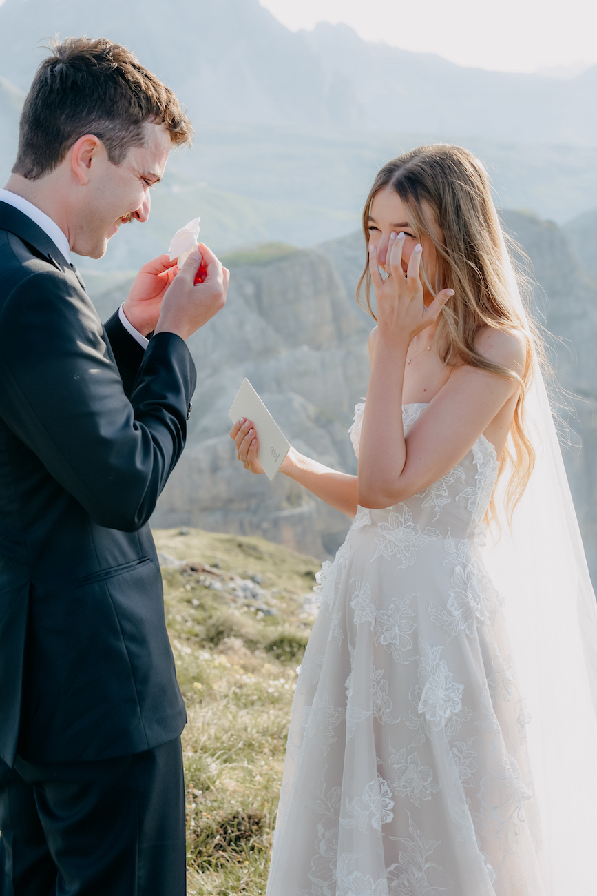A bride and groom stand outdoors on a grassy hill, smiling and wiping tears while exchanging handwritten notes on their wedding day.