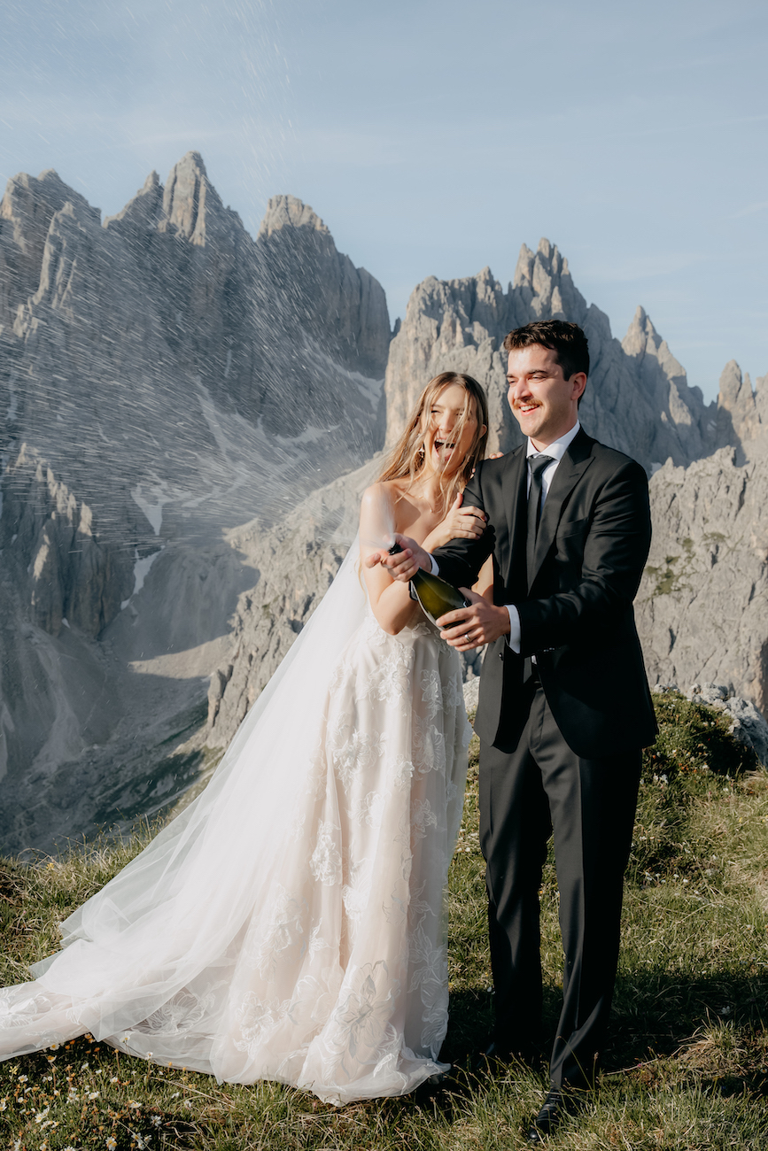 A bride in a white gown and a groom in a black suit open a champagne bottle together outdoors with rocky mountains in the background.