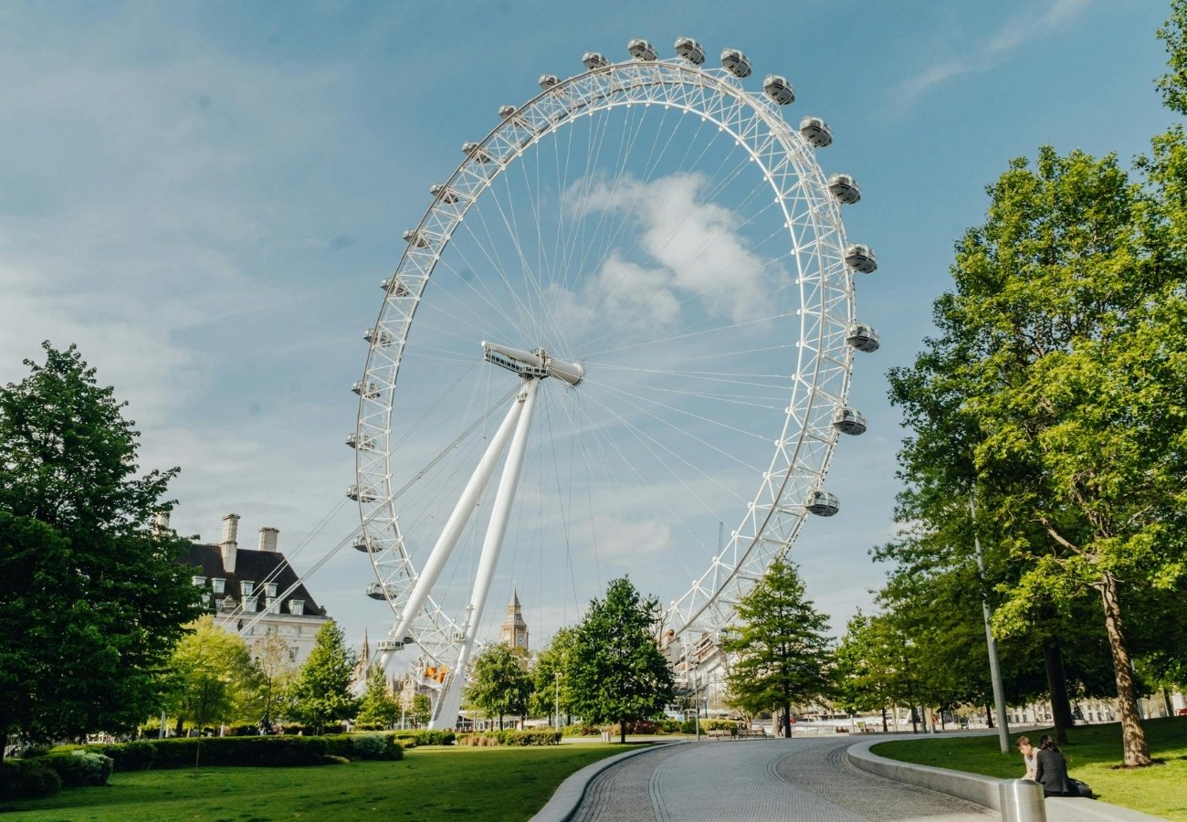 A large Ferris wheel, known as the London Eye, stands by a curved pathway surrounded by green trees and grass under a partly cloudy sky.