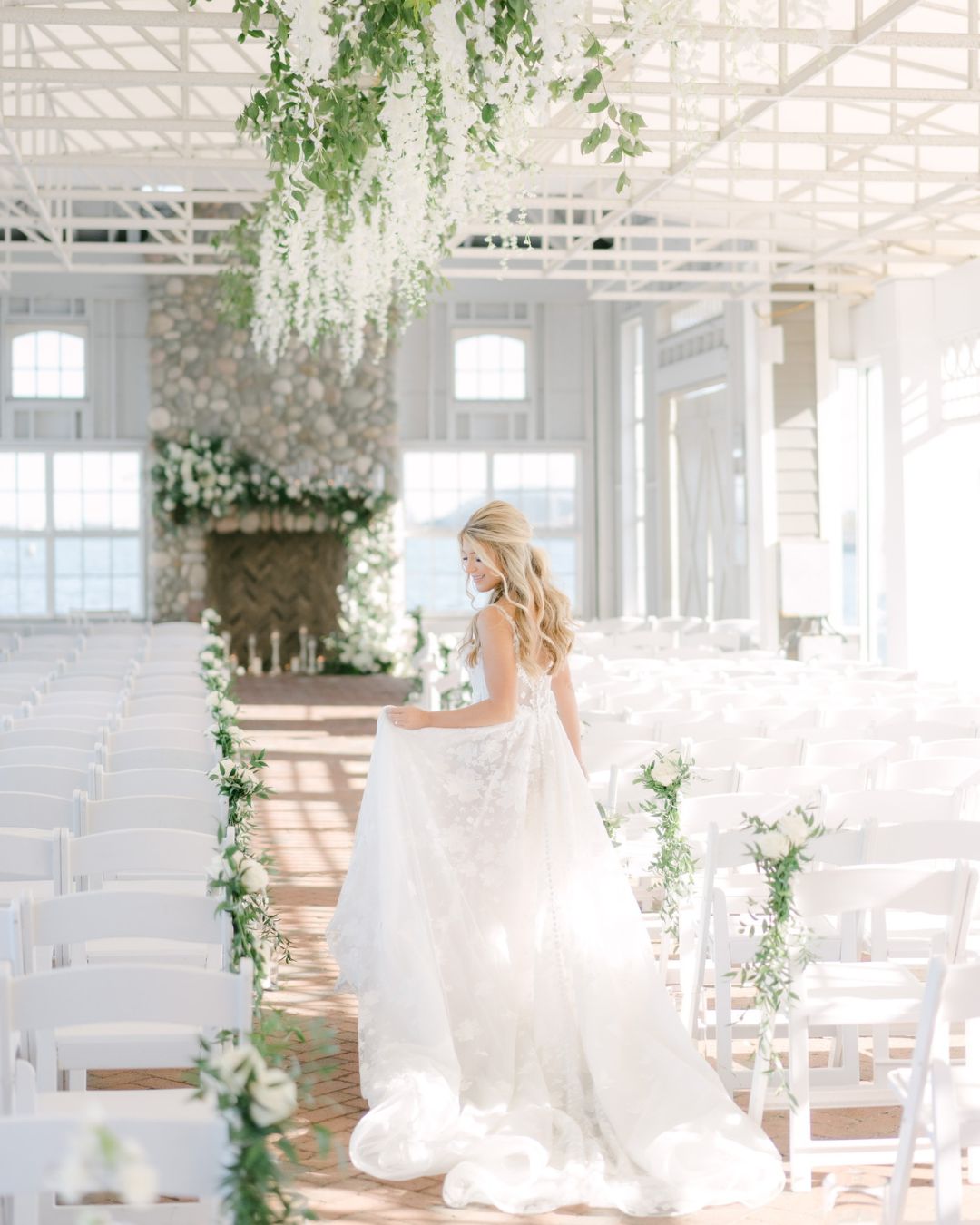 Bride in a white wedding dress walks down an aisle lined with white chairs and greenery in a bright, airy venue with large windows and floral decorations.