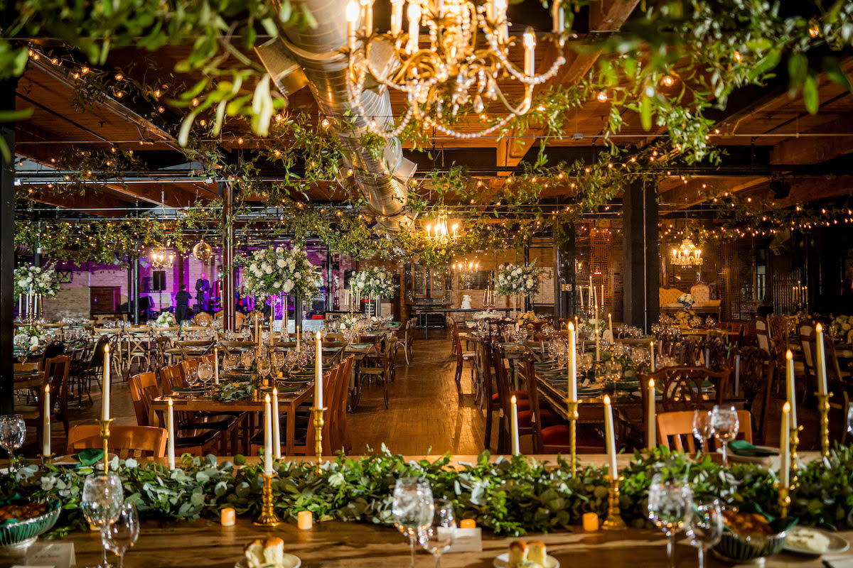 A decorated banquet hall features chandeliers, string lights, greenery, long tables with candles, floral centerpieces, and elegant place settings.