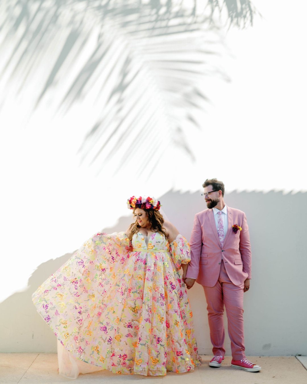 A woman in a floral dress and flower crown stands next to a man in a pink suit against a white wall with palm shadows.