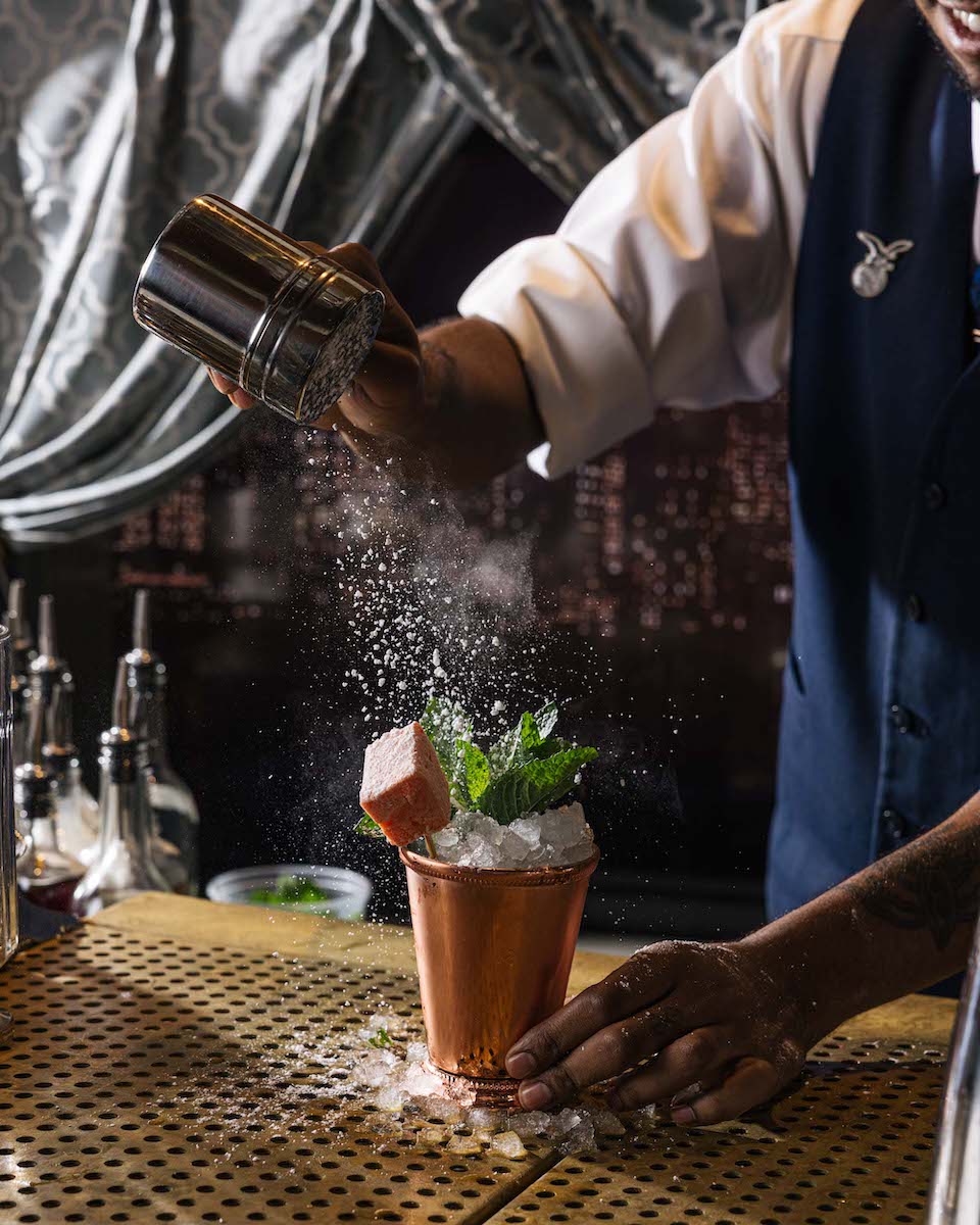 A bartender sprinkles powdered sugar over a cocktail garnished with mint in a copper cup on a bar counter.