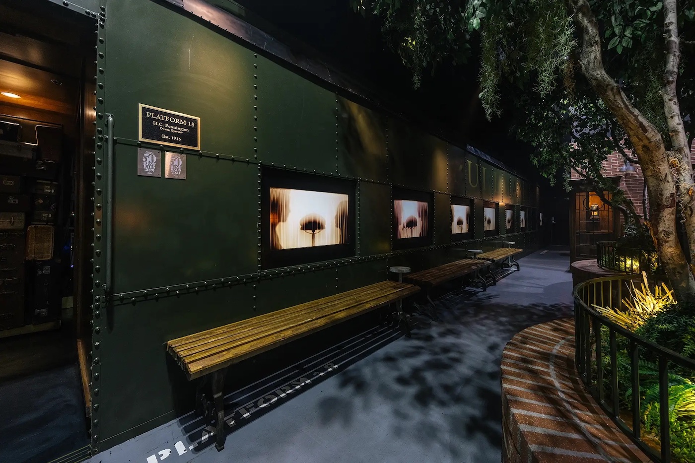 A dimly lit hallway with green walls features several illuminated display screens and wooden benches along one side, with trees and plants visible nearby.
