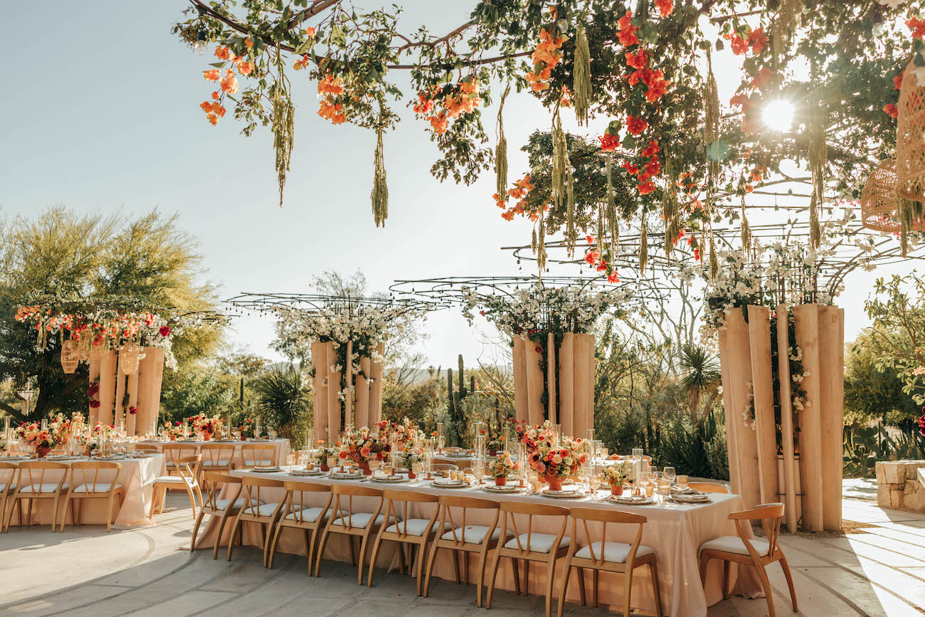 Outdoor wedding reception setup with long tables, floral centerpieces, hanging flowers, and sunlight streaming through greenery.