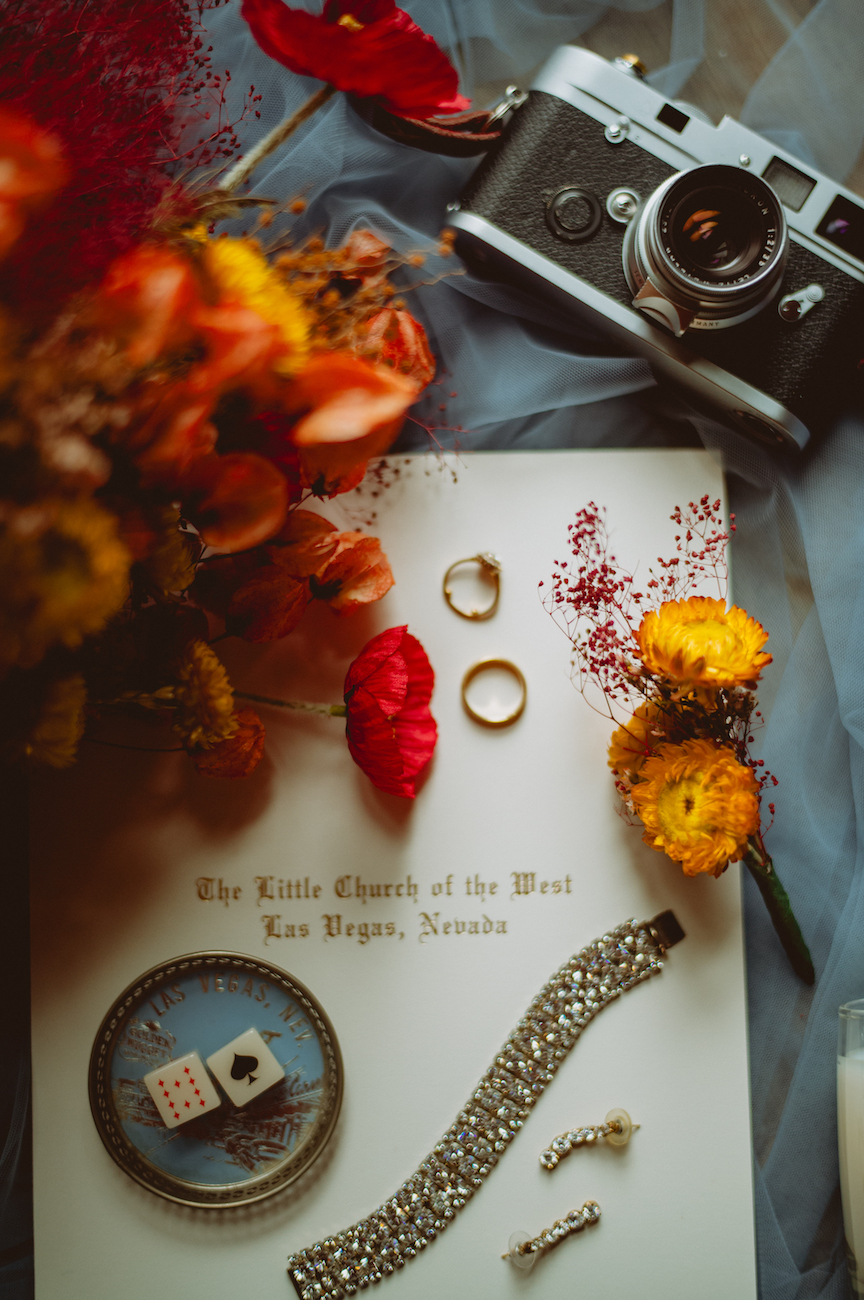 Wedding-themed flat lay featuring flowers, two gold rings, a vintage camera, jewelry, and a booklet titled “The Little Church of the West, Las Vegas, Nevada.”.