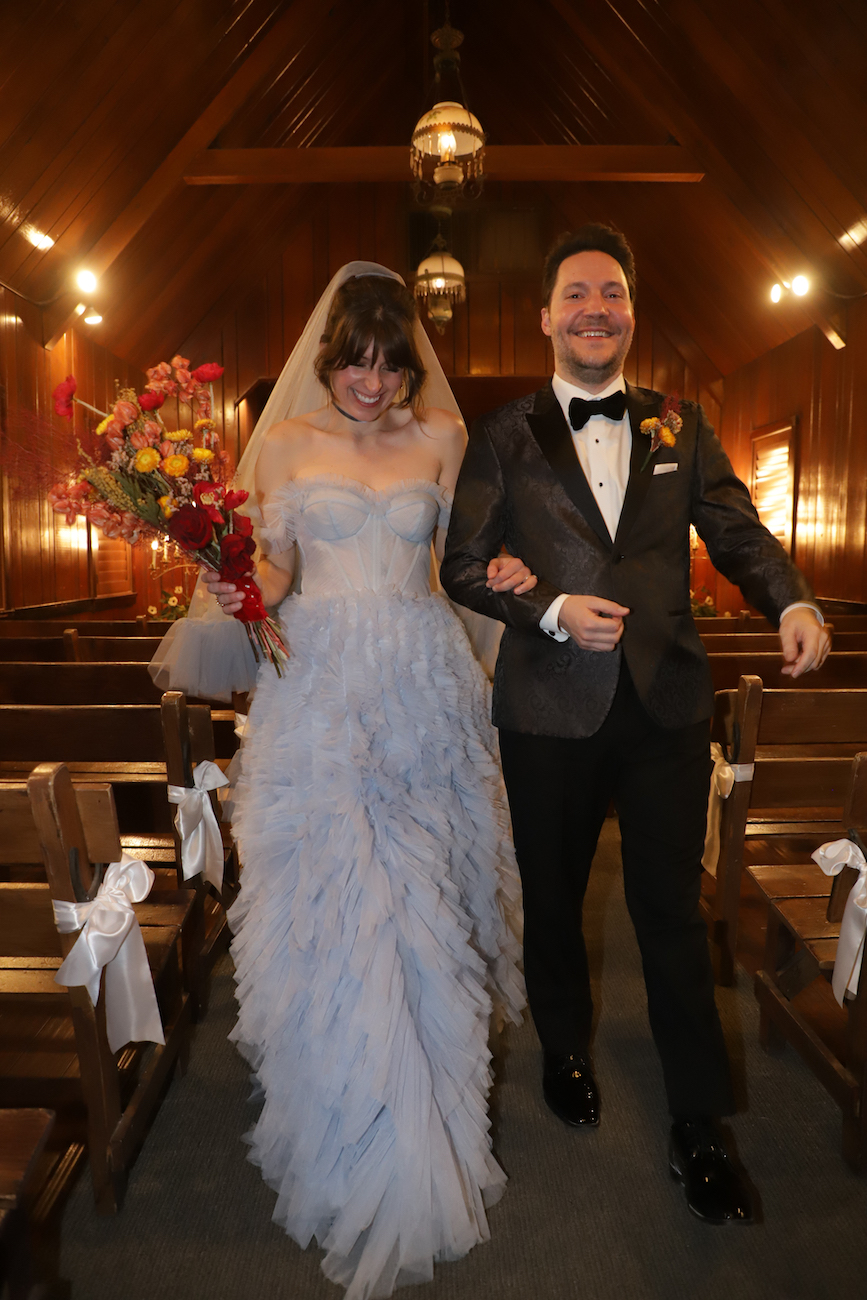 A bride in a light blue strapless gown and a groom in a black tuxedo walk arm in arm down the aisle of a dimly lit wooden chapel.