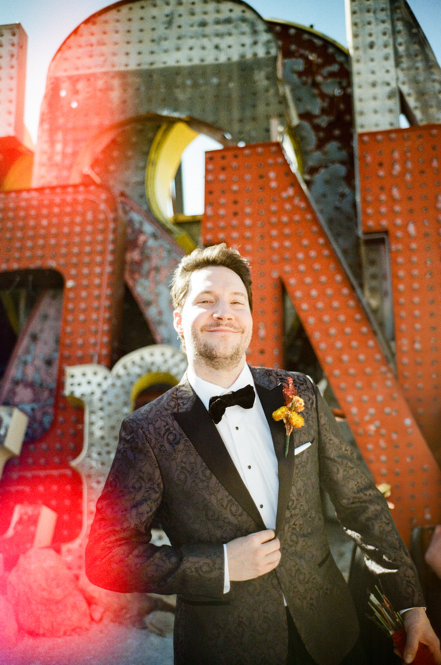 Man in a black tuxedo with a bow tie and boutonniere stands smiling in front of large, vintage marquee letters in daylight.