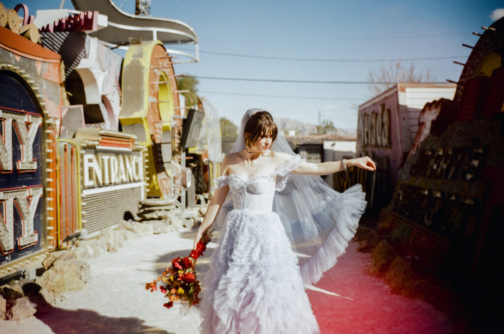 A bride in a white ruffled dress and veil stands outdoors holding a bouquet of red flowers, beside vintage neon signs under a clear sky.