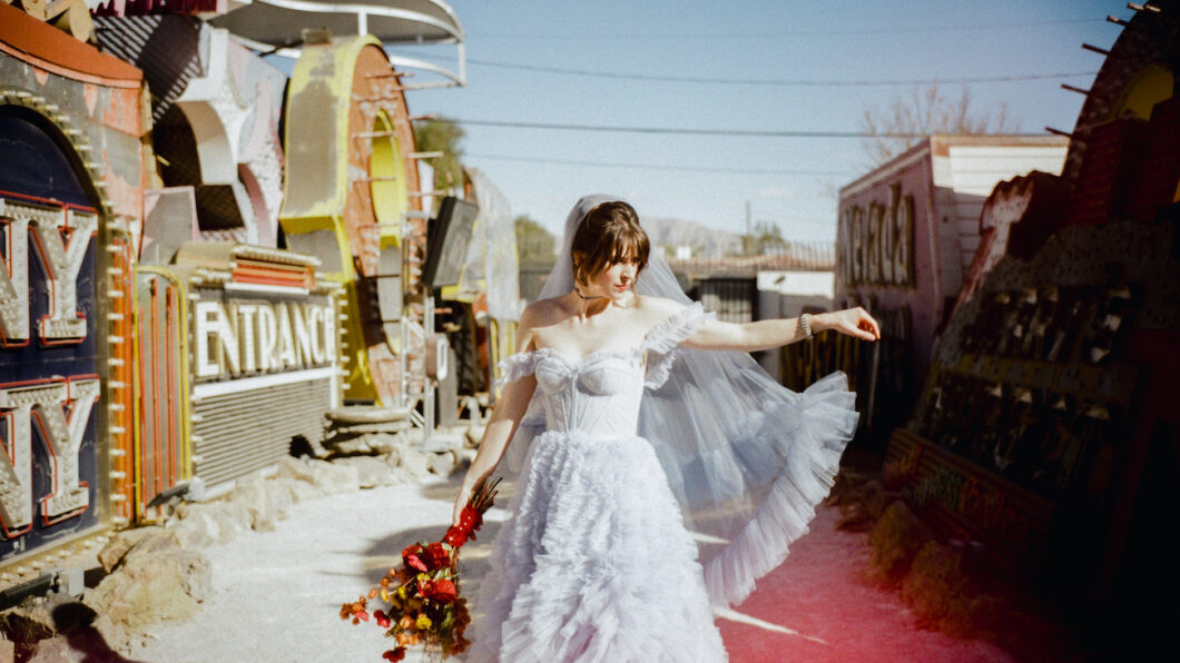 A bride in a white ruffled dress and veil stands outdoors holding a bouquet of red flowers, beside vintage neon signs under a clear sky.
