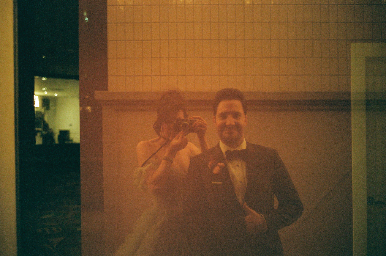 A man in a suit and a person with a camera are reflected in a glass surface; the image is tinted with a warm, yellowish hue.