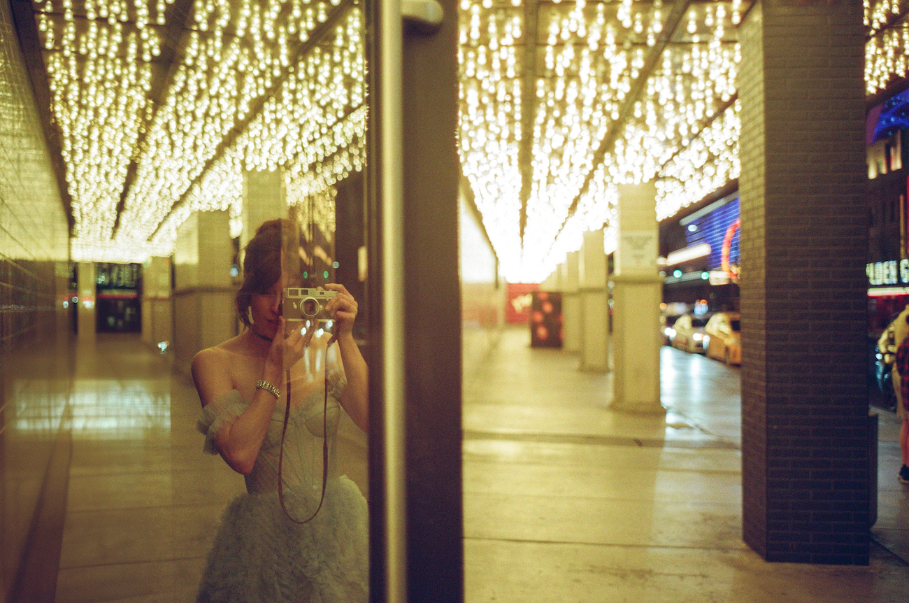 A person takes a photo of their reflection in a glass wall under a ceiling of bright string lights on a city street at night.