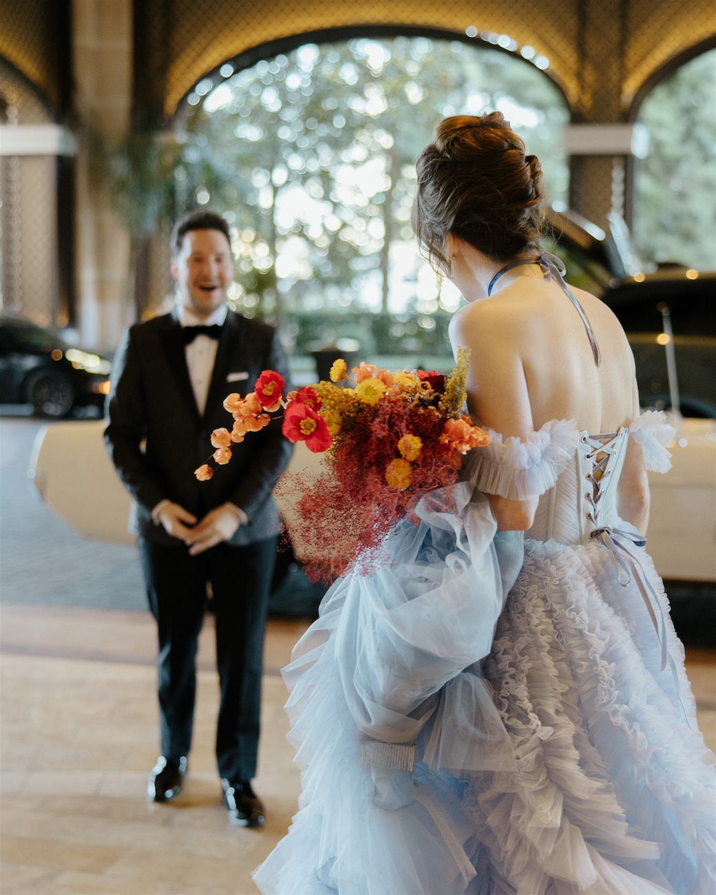 A person in a tuxedo stands smiling with surprise as another person in a light blue gown holding a colorful bouquet approaches them indoors.