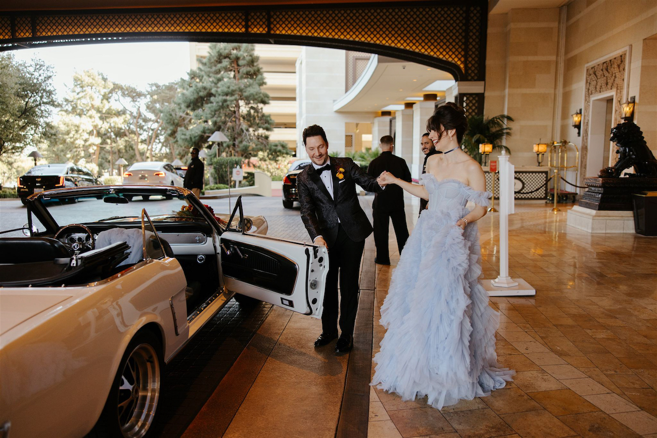 A man in a tuxedo holds open the door of a white convertible for a woman in a light blue gown outside a hotel entrance.