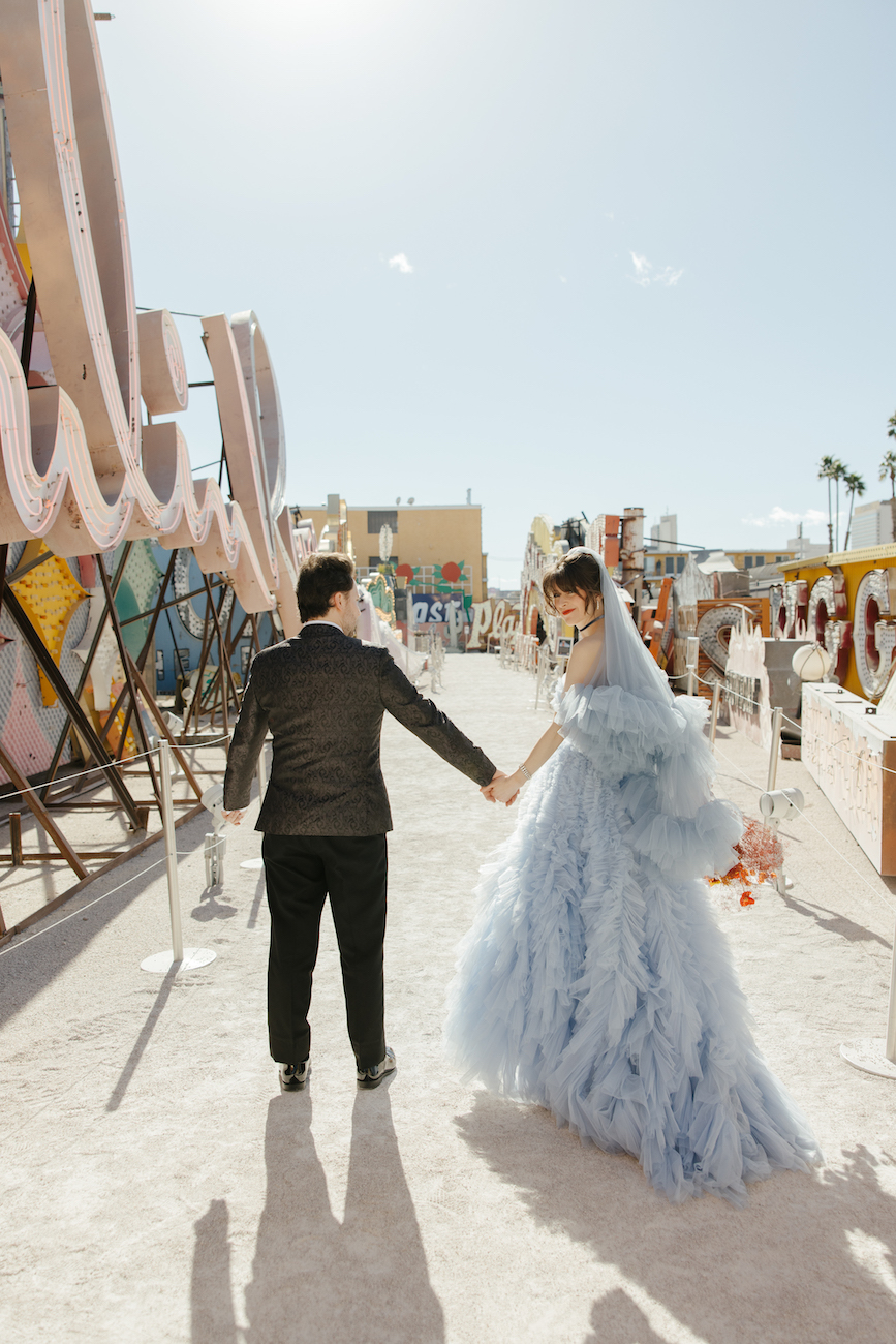 A couple holding hands walks outdoors; the person on the left wears a black suit, and the person on the right wears a light blue, ruffled dress and veil.