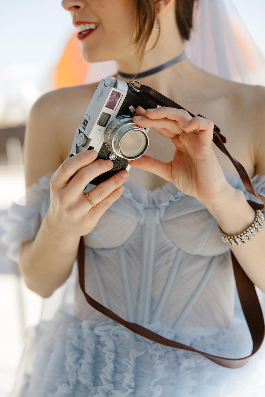 Woman in a light blue dress holding a vintage camera, with a veil and bracelet visible, photographed from the shoulders down.