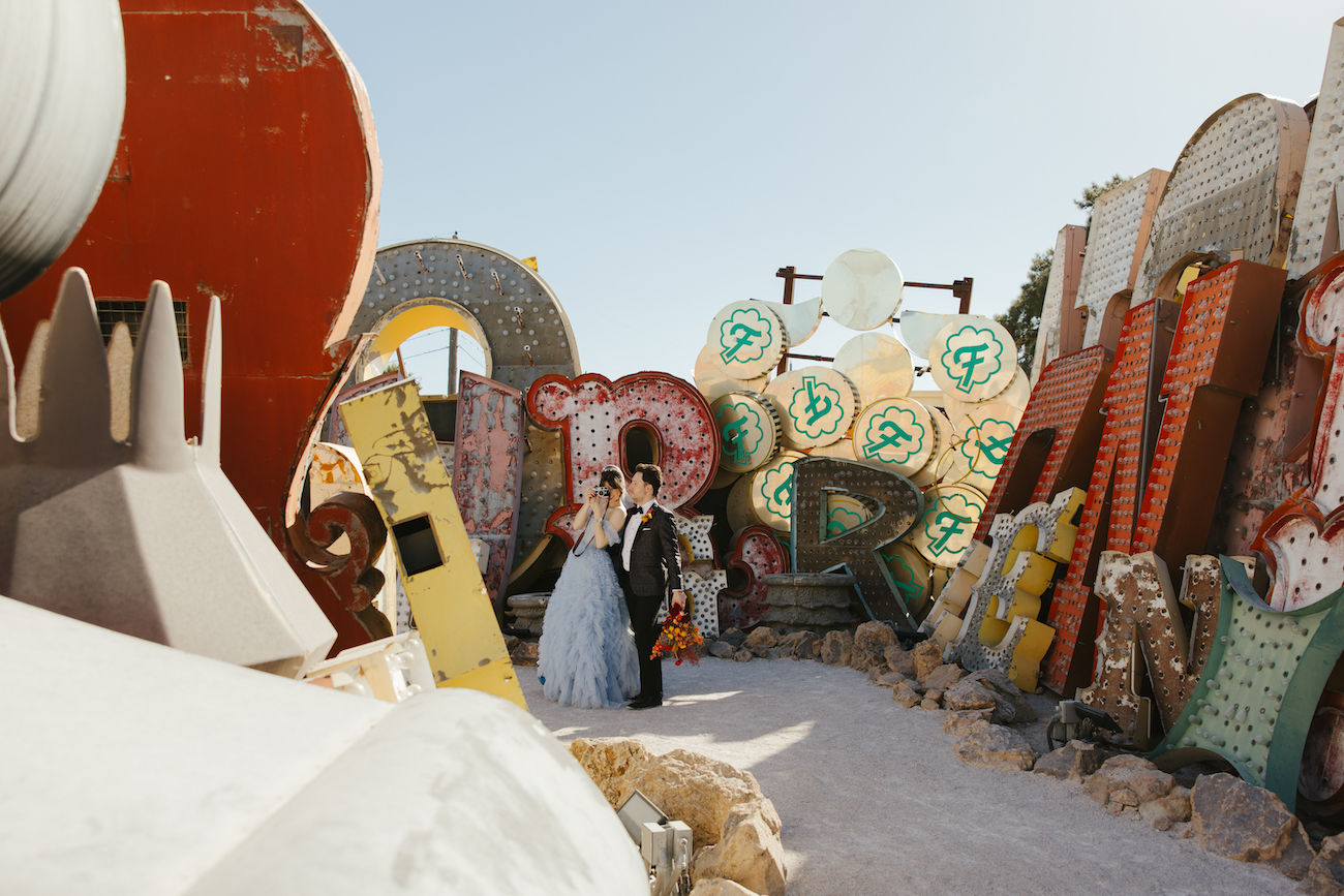 A couple poses together near colorful, vintage neon signs in an outdoor setting on a sunny day.