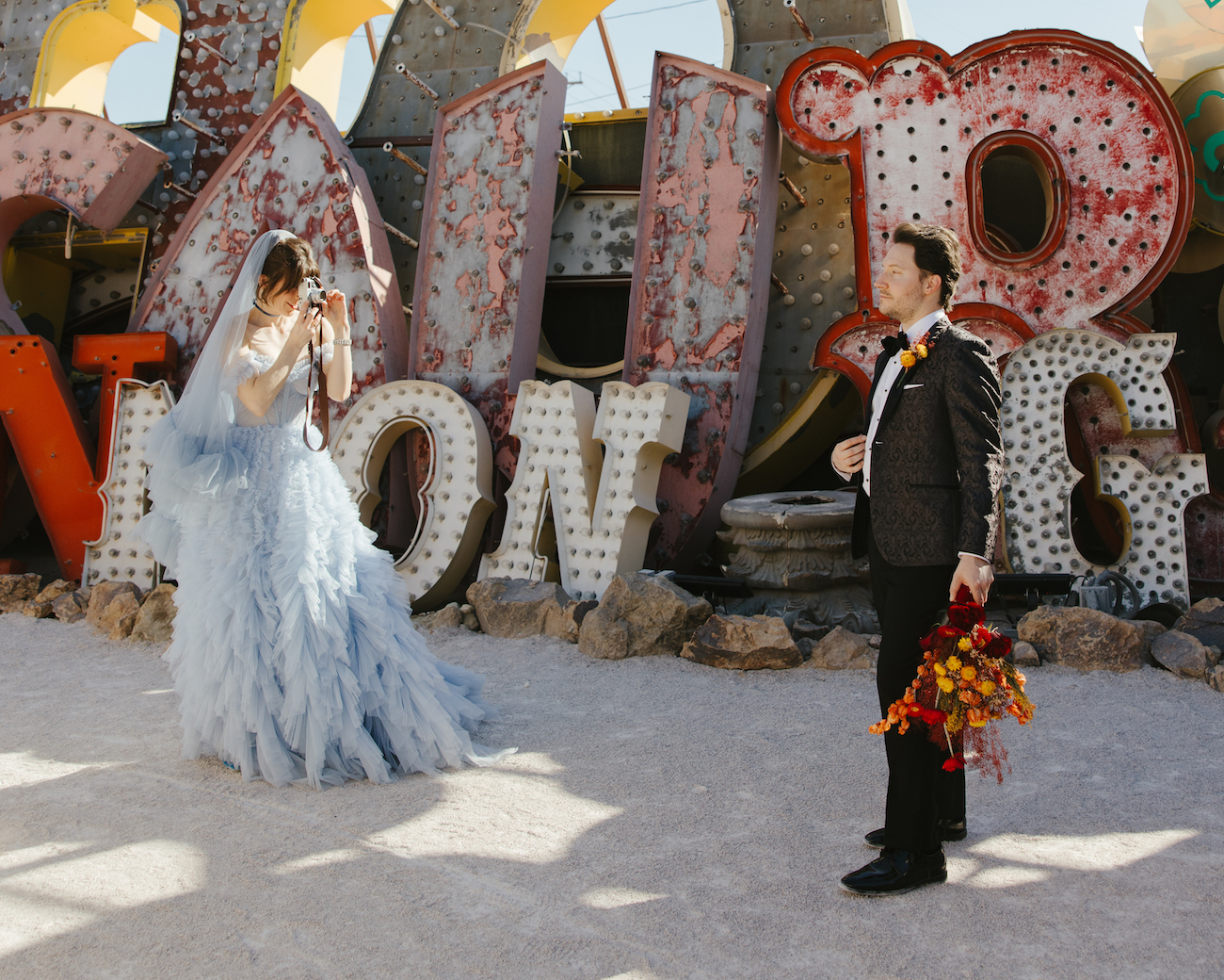 A bride in a blue dress takes a photo of a groom holding a bouquet in front of large, vintage marquee letters outdoors.