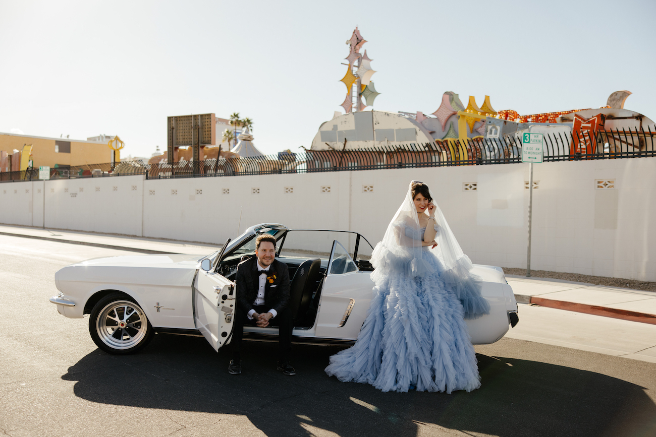 A couple dressed in formal attire poses with a white classic convertible parked on a street, with colorful retro signs visible in the background.