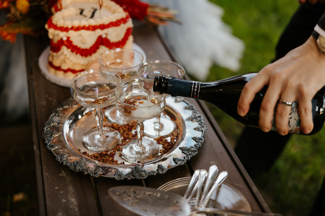 A person pours sparkling wine into coupe glasses on a silver tray next to a decorated cake and some silverware on a wooden table.