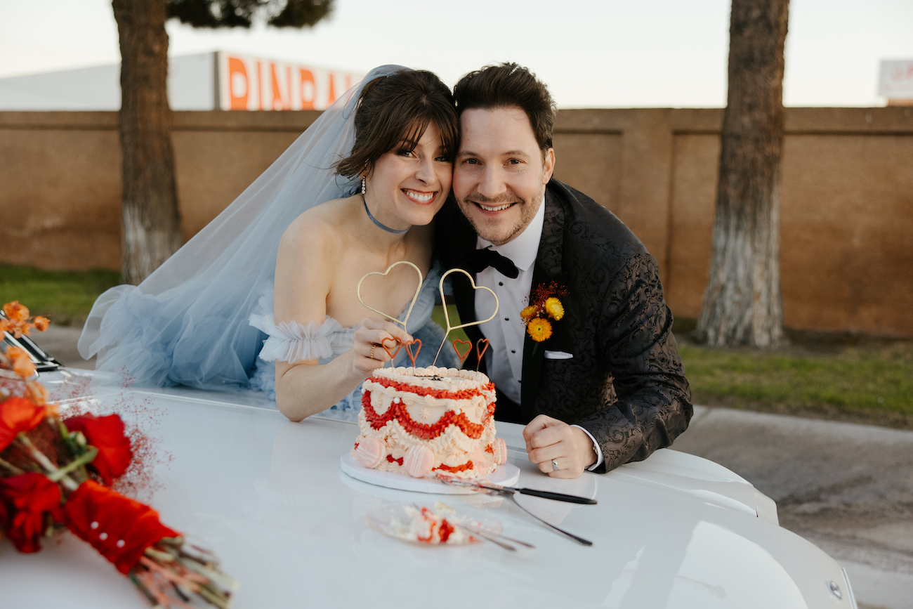 A bride and groom in wedding attire smile while posing with a small decorated cake on the hood of a white car.
