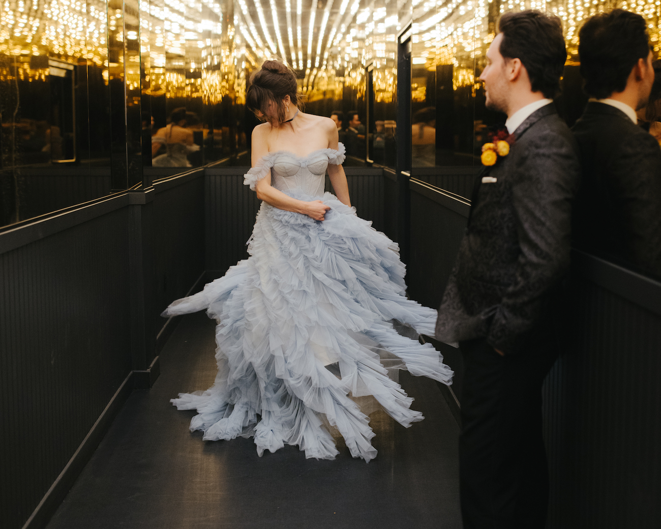 A woman in a light blue ruffled gown spins while a man in a dark suit with a yellow boutonniere watches in a hallway with mirrored walls and lights on the ceiling.