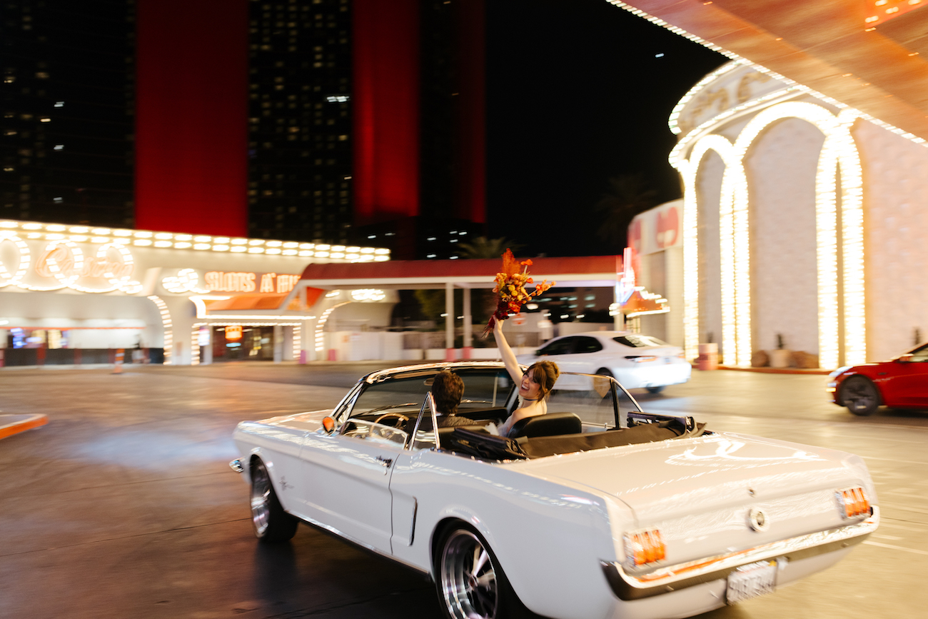A person in a white convertible holds a bouquet of flowers while driving past brightly lit buildings at night.
