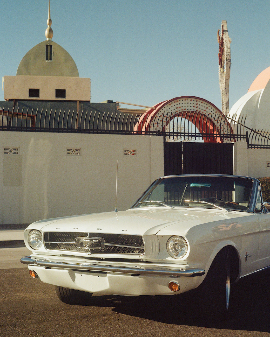 A white classic Ford Mustang convertible parked in front of a modern building with domes and decorative arches under a clear sky.