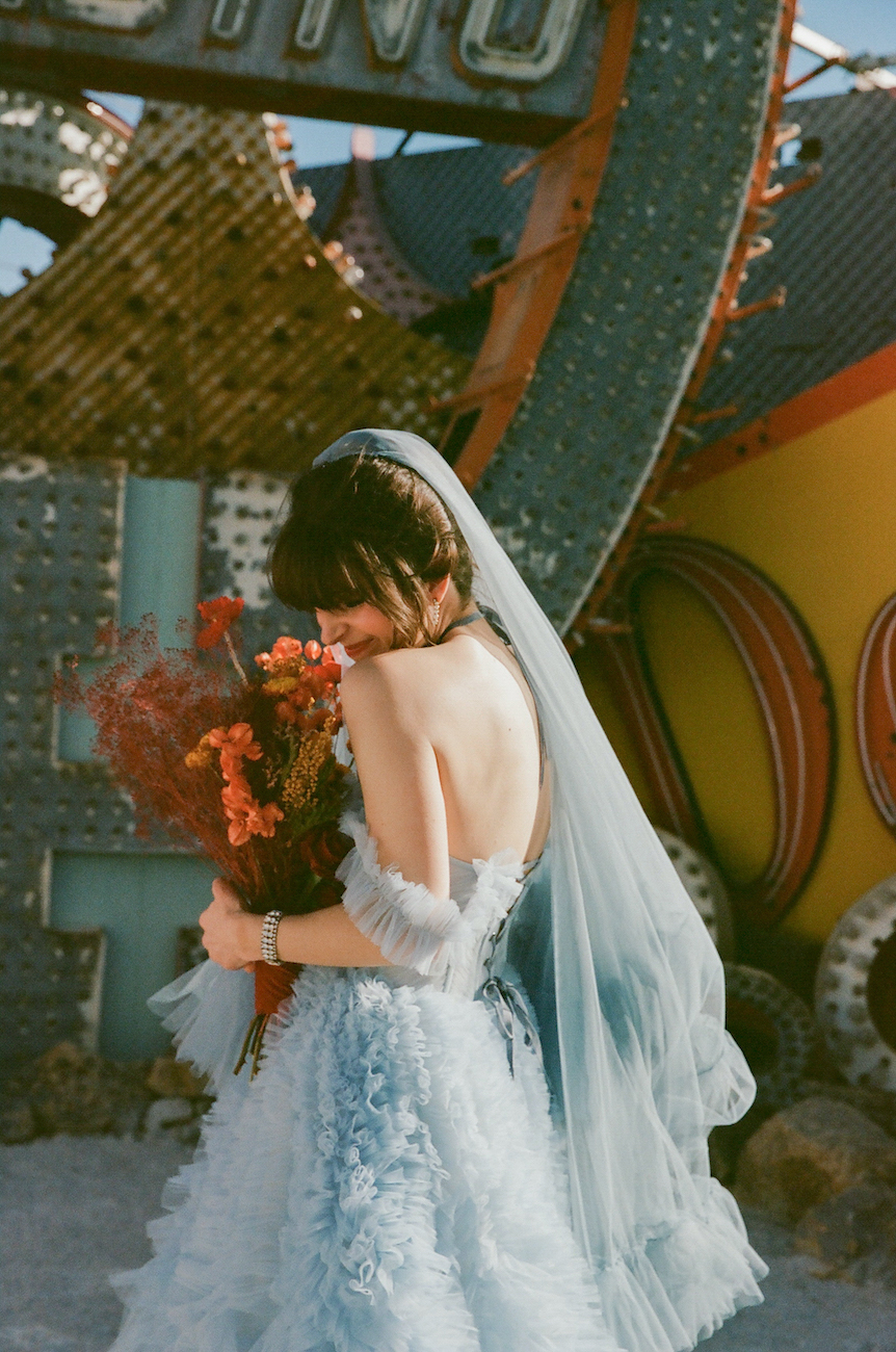 A bride in a light blue gown and veil holds a bouquet of red flowers, standing outdoors in front of vintage neon signs.