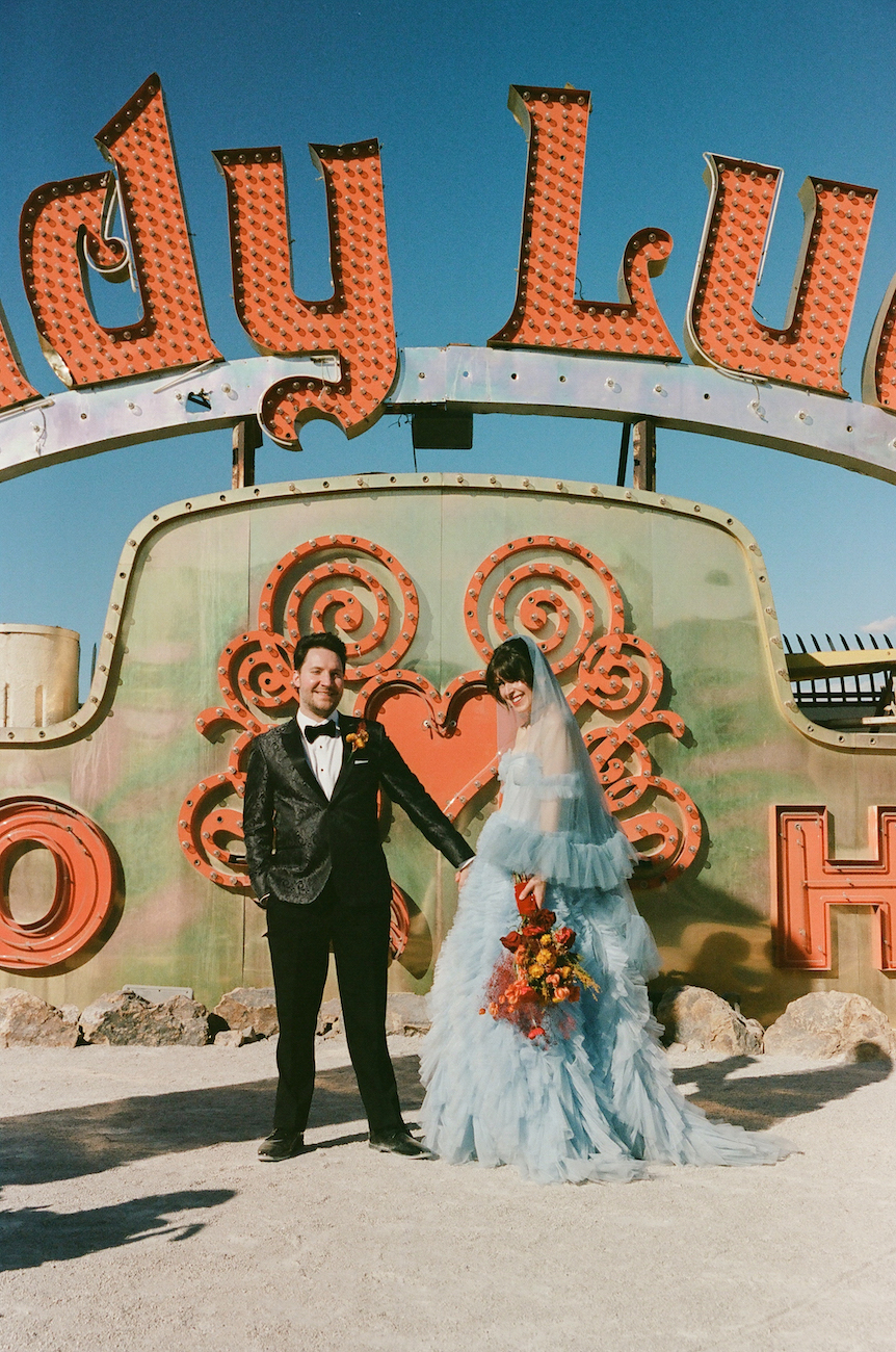 A couple in formal attire stands holding hands in front of a vintage "Lady Luck" sign, with the woman holding a bouquet and wearing a blue dress and veil.
