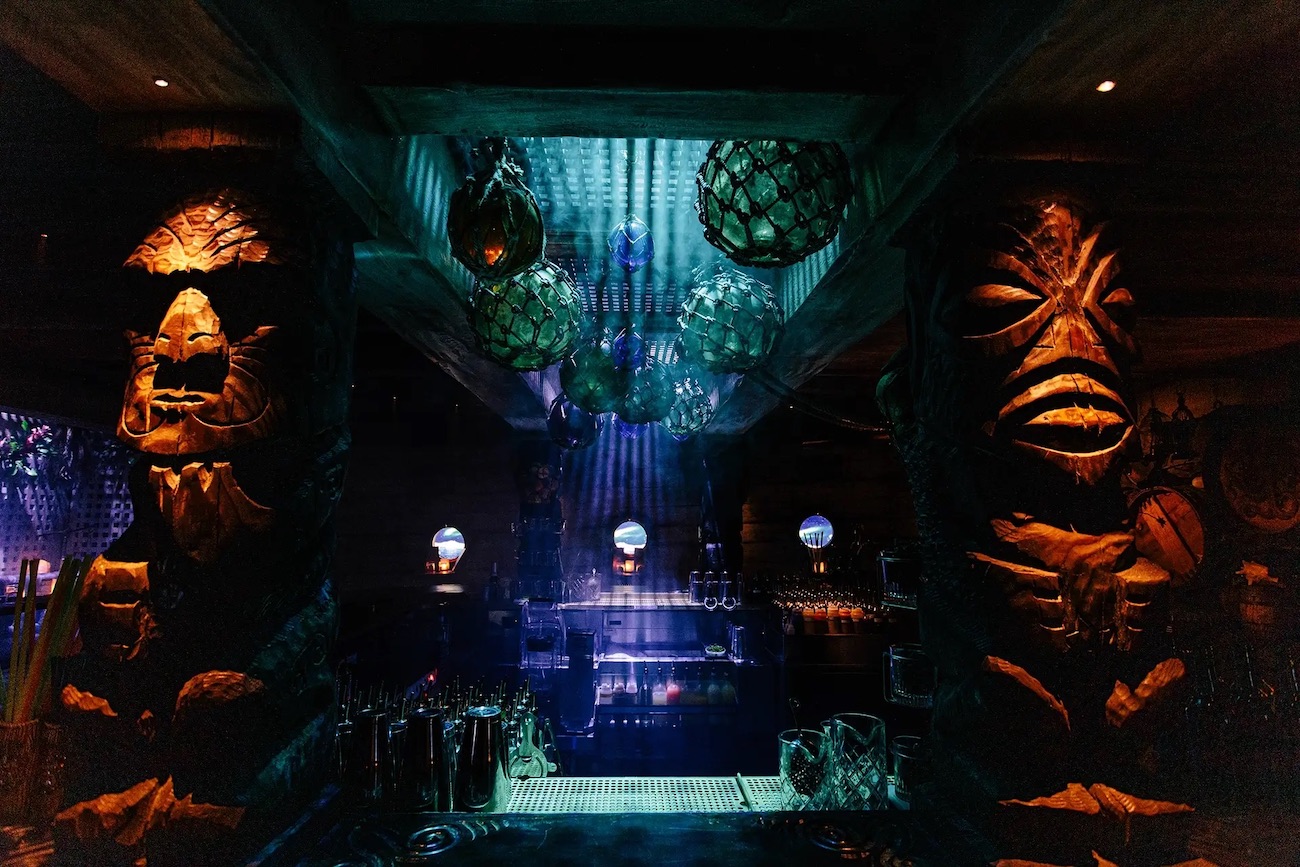 Dimly lit bar with tiki statues, hanging lanterns, and blue-purple ambient lighting, creating a tropical, exotic atmosphere.