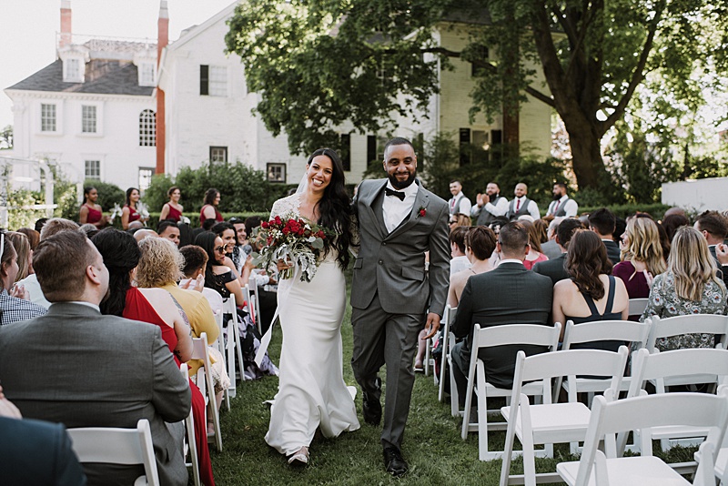 A bride and groom walk down the aisle outdoors, smiling, with guests seated on both sides and a large white house in the background.