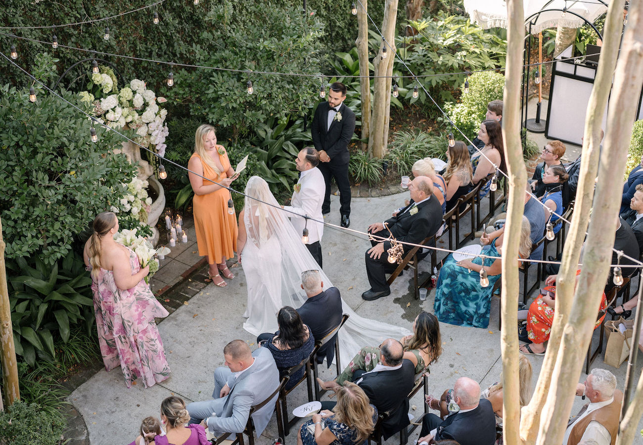 Outdoor wedding ceremony seen from above, with a bride and groom standing together, officiant in front, and guests seated on either side surrounded by greenery and string lights.