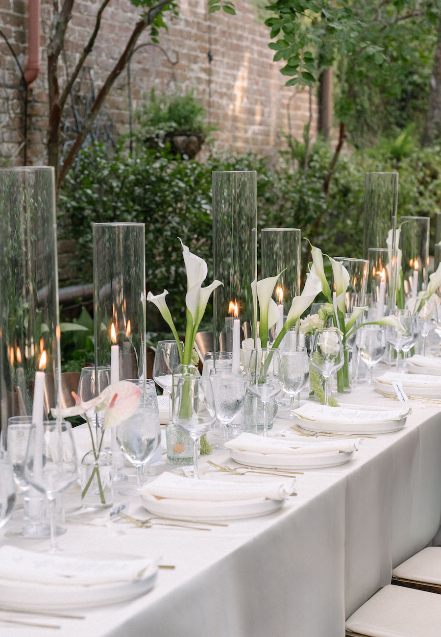 A long outdoor dining table is set with white plates, glassware, tall glass candle holders, and vases with white calla lilies.