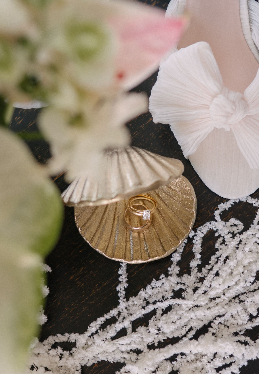 A gold ring in a shell-shaped jewelry box, surrounded by white beaded accessories and a pale pink shoe with a bow.