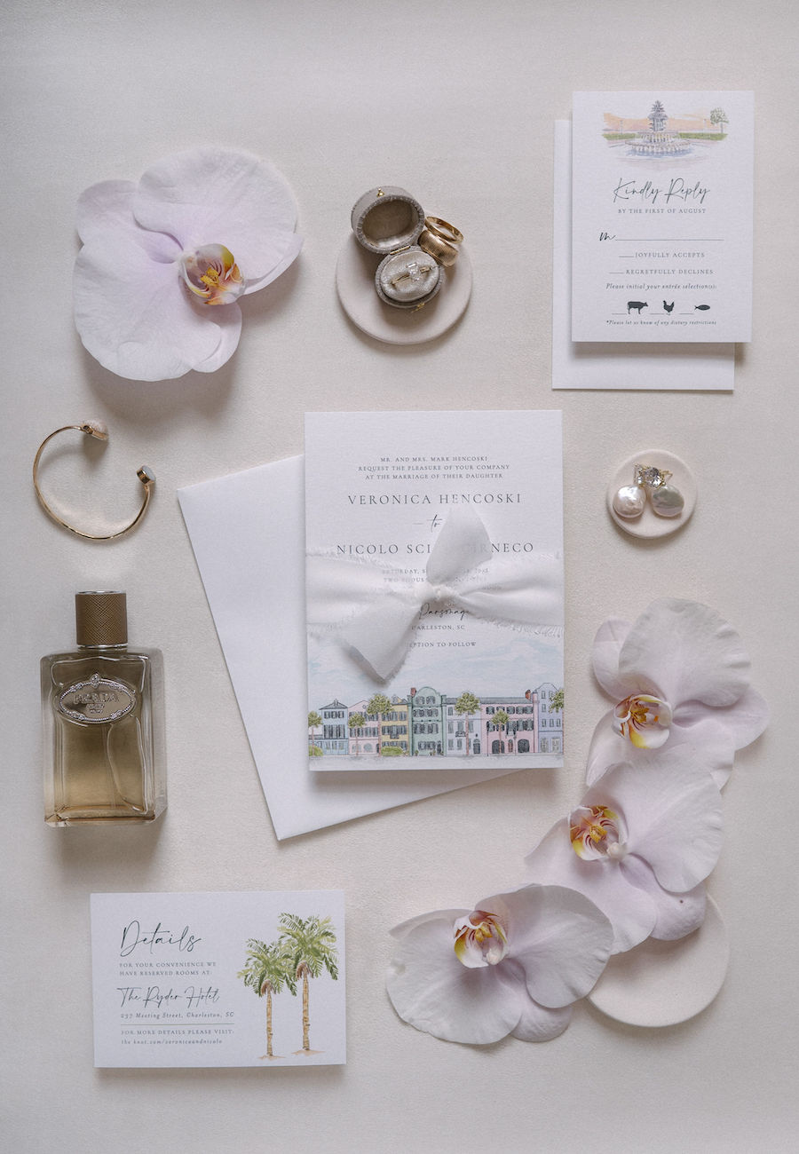 Flat lay of wedding stationery, white orchids, perfume bottle, rings, a bracelet, and a small dish on a light surface.