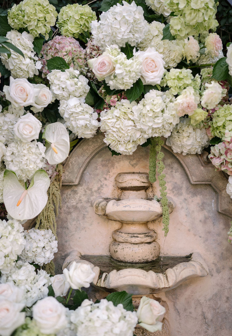 Stone fountain surrounded by white hydrangeas, roses, and green foliage against a wall.