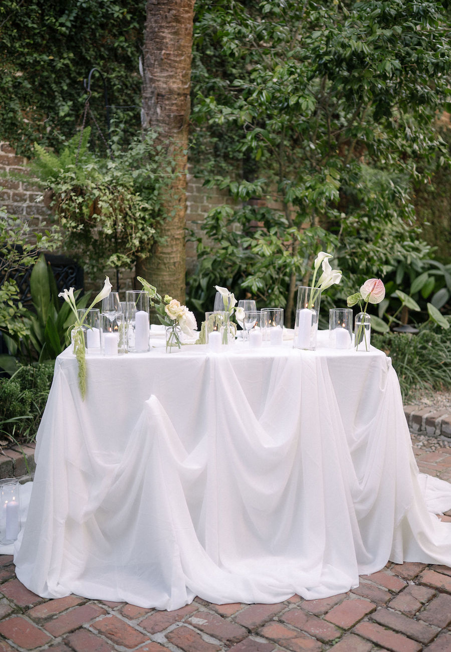 A round table covered with a white draped tablecloth, decorated with white flowers and candles, set outdoors on a brick patio with greenery in the background.