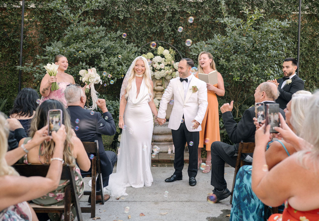 A bride and groom stand hand-in-hand at an outdoor wedding ceremony, surrounded by guests taking photos and members of the wedding party.