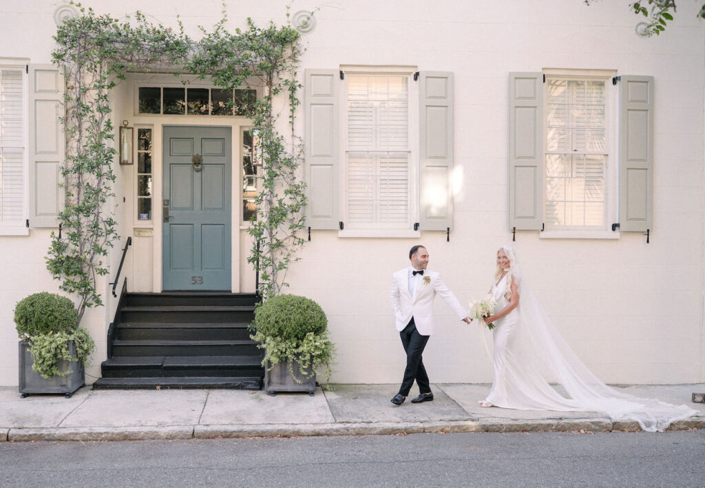 A bride and groom stand hand in hand on a sidewalk in front of a light-colored building with green shutters and a green door.
