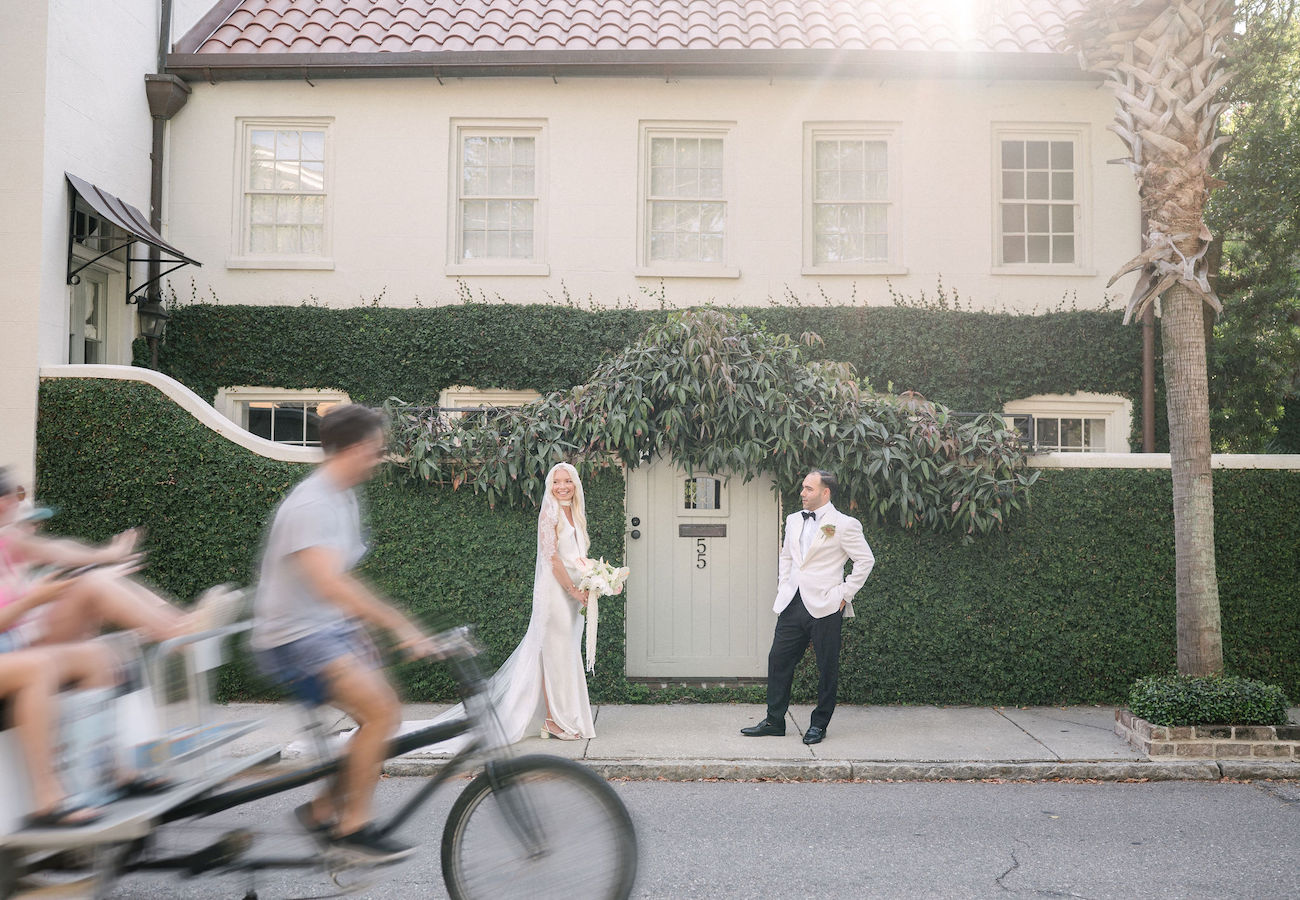 Bride and groom pose on a sidewalk outside a house while a cyclist pulls a passenger in the foreground, slightly blurred.