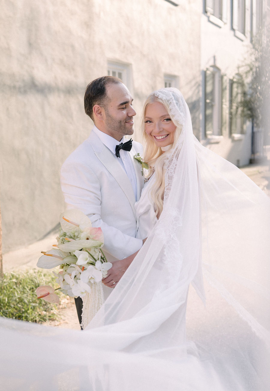 A bride in a white lace veil and a groom in a white jacket and black bow tie stand together outdoors, smiling and holding a bouquet of white flowers.