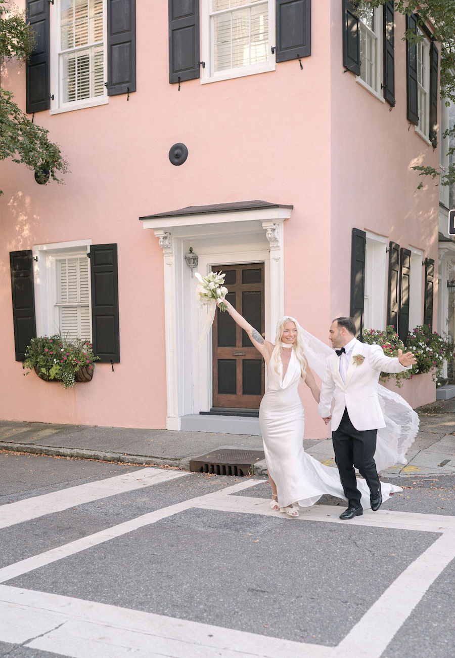 A bride and groom cross a street holding hands, with the bride raising her bouquet, in front of a pink building with black shutters.
