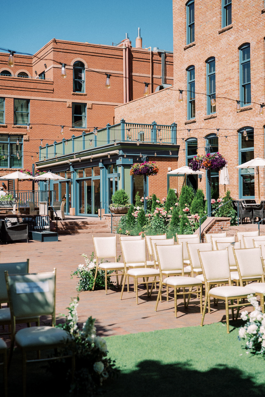 Outdoor wedding ceremony setup with rows of white chairs on a patio, surrounded by brick buildings and greenery, under a clear blue sky.