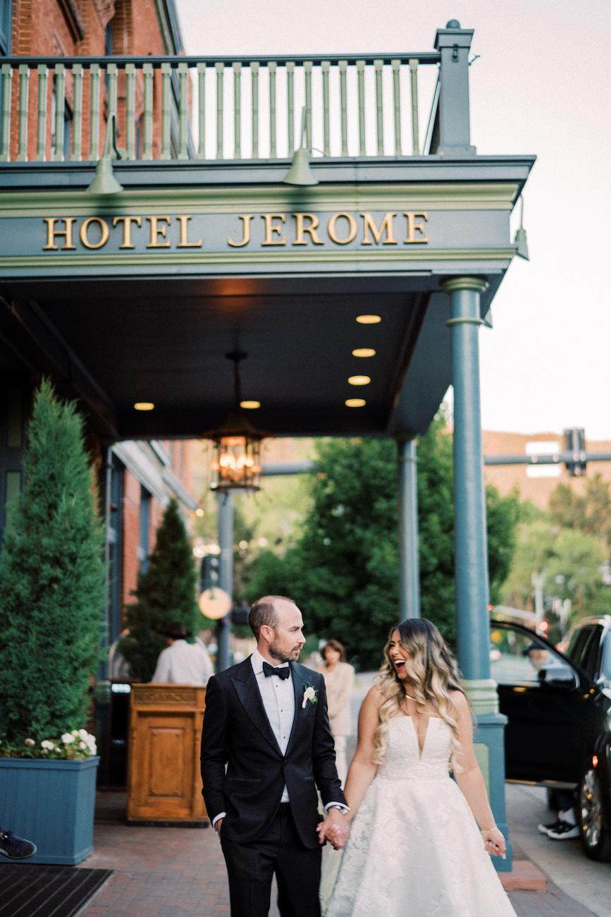 A bride and groom hold hands and walk outside the entrance of Hotel Jerome, dressed in formal wedding attire.