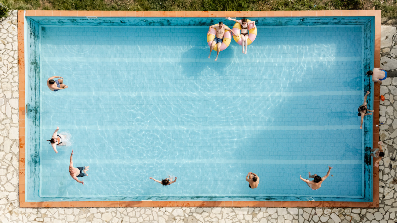 Aerial view of a rectangular swimming pool with several people swimming and three people sitting on pool floats near one edge.