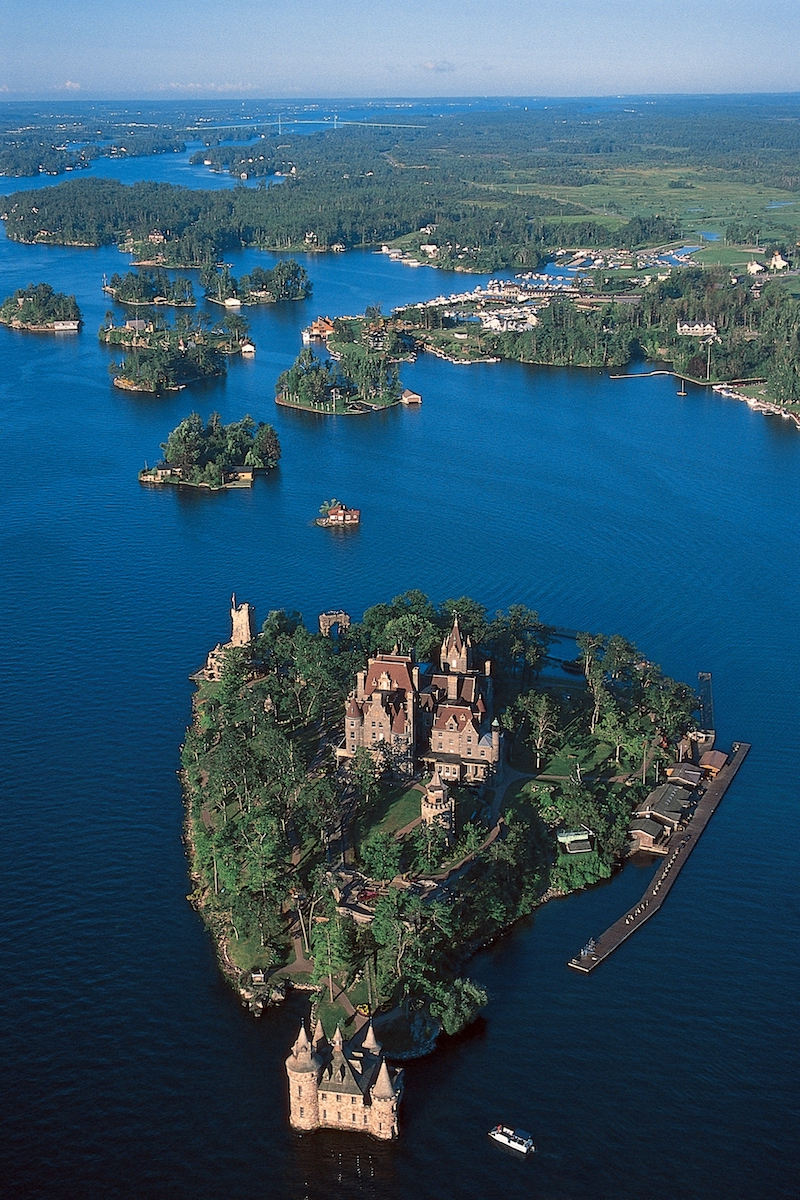 Aerial view of a forested island with a large historic castle surrounded by water and smaller islands, with a boat docked nearby.